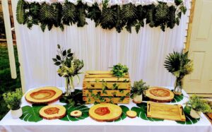 DRAPED BACKDROP WITH ARTIFICIAL GREENERY VALANCE AND DESSERT TABLE TABLE SETUP WITH SERVEWARE AND TABLE DECOR